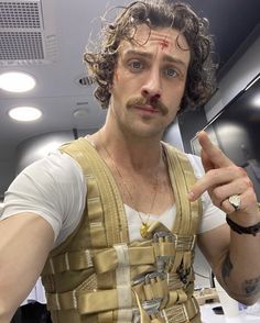 a man with long curly hair wearing a vest and pointing to the side while standing in front of a mirror