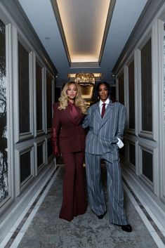 two women in business attire standing next to each other on a long hallway with chandeliers