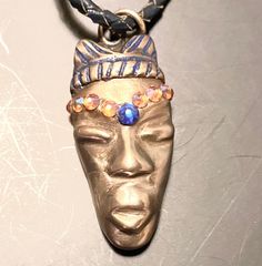 AC5. Handmade bronze pendant African Queen, with adornments, 8 Swarovski Crystals that change color as they catch the light, from an orange rose color, to blue.  The center stone is also a Swarovski Crystal that reflects the light and changes from navy blue to violet rose.   Additionally,the crown has been enhanced with a blue jewelers dye.  Very striking.  The Queen is hung from a cord that has been made by braiding navy color leather. This Queen differs from the Queen made of sand bronze, as t Spiritual Gold Jewelry With Waxed Cord, Artisan Gold Jewelry With Adjustable Cord, Copper Necklace With Adjustable Cord As Gift, Unique Waxed Cord Pendant Jewelry, Adjustable Bronze Hand Cast Necklace, Artisan Gold Necklace With Adjustable Cord, Rose Violette, Rose Orange, African Queen