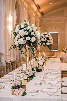 a long table with white and pink flowers on it, surrounded by gold place settings