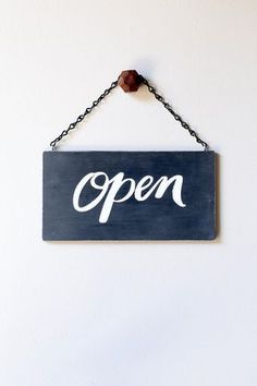 an open sign hanging from a chain on a white wall with a wooden bead