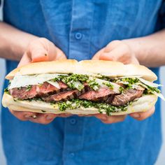 a person holding a sandwich with meat and greens on it in their hands while wearing a blue shirt