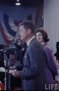 a man standing in front of a microphone next to two women and an american flag