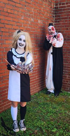 two people dressed as clowns are standing next to a brick wall with grass in front of them