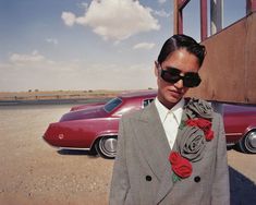 a man in a suit and sunglasses standing next to a red car on the road