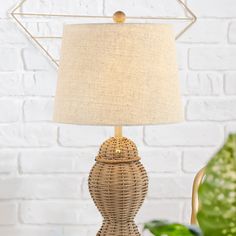 a wicker table lamp with a white shade on it and a green plant in the background