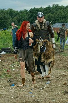 a man and woman walking next to a goat on a dirt field with other people in the background