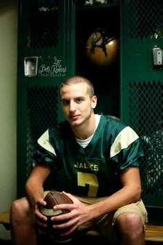 a football player sitting in the locker room holding a ball and posing for a photo