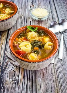 two bowls of tortellini soup on a wooden table with silverware and spoons