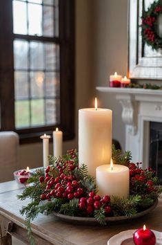 candles are lit on a tray with holly and berries in front of a fire place