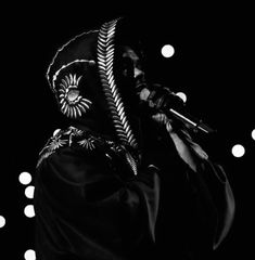 a man with a hood on holding a microphone in front of his face and lights behind him