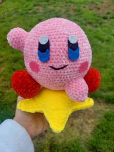 a hand holding a pink stuffed animal with blue eyes and a yellow star in the grass