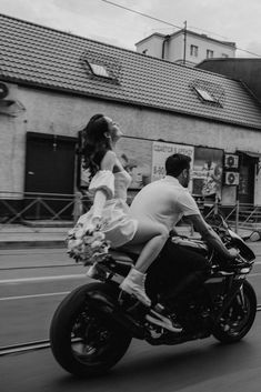 a man and woman riding on the back of a motorcycle down a street with buildings in the background