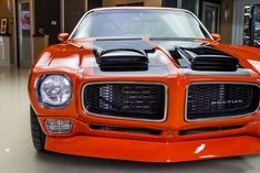 an orange sports car parked in a showroom with its hood up and lights on