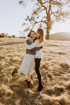 two women hugging each other in the middle of a field with a tree behind them