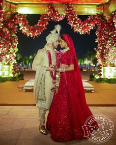 two people standing next to each other in front of a decorated archway with red flowers