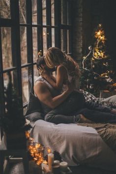 a man and woman sitting on a bed in front of a window with christmas trees