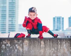 a little boy dressed as spider man sitting on top of a cement wall with his hands in the air