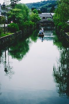 two people in a small boat traveling down a narrow river surrounded by trees and houses