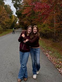 two young women hugging each other while standing on the side of a road with trees in the background