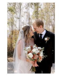 a bride and groom standing together in front of trees with their wedding bouquets on