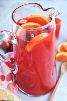 a pitcher filled with red liquid and sliced oranges next to other fruit on a table