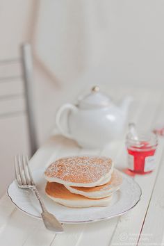 two pancakes on a plate next to a teapot and knife, with sugar in the foreground