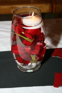 a glass filled with water and red flowers next to a lit candle on top of a table