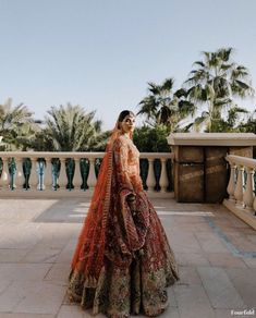 a woman in an orange bridal gown stands on a balcony with palm trees behind her