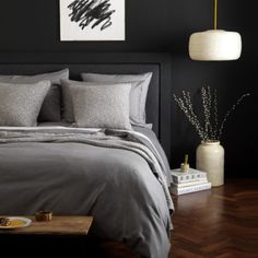 a bed with grey linens and pillows in a black walled room, next to a painting on the wall