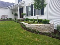 a stone wall in front of a white house with green grass and bushes around it