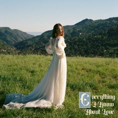 a woman in a long white dress standing on top of a lush green field with mountains in the background