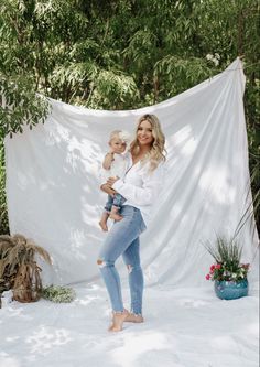 a woman holding a baby standing in front of a white sheet with trees behind her