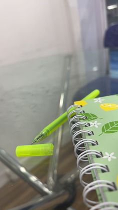 a spiral notebook with two green pens on it and a clear plastic container in the background