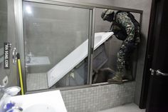 a man in camouflage standing next to a bathtub and sink with the door open