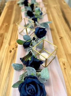 blue roses and greenery are arranged on a table runner with gold geometric vases