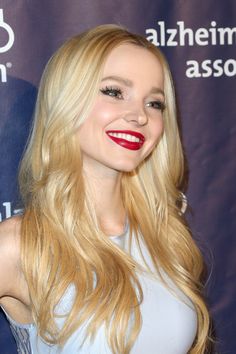 a woman with long blonde hair and bright red lipstick smiles at the camera while standing in front of a blue backdrop