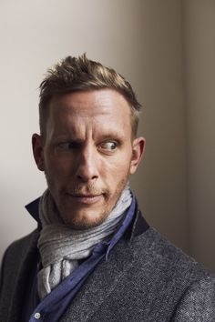 a man wearing a gray jacket and blue scarf posing for a photo in front of a white wall