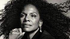 a black and white photo of a woman with curly hair looking at the camera while holding her hand on her chin