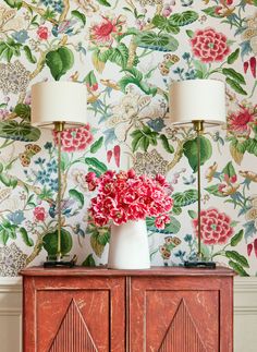 two lamps on top of a wooden cabinet in front of a floral wallpaper