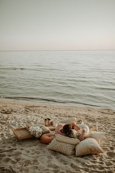 a woman laying on top of a beach next to the ocean with pillows and food