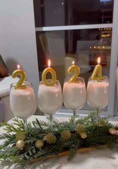 three glasses filled with white liquid and gold numbers on top of a table covered in greenery