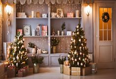 two christmas trees in front of a book shelf with presents on it and lights around them