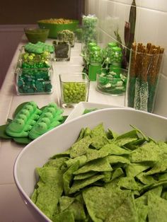 a bowl filled with green chips next to other snacks on a white counter top in front of bottles and glasses