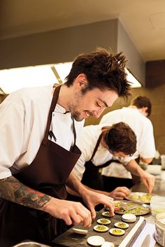 two men in aprons are preparing food on a counter top with other chefs behind them