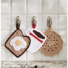 three crocheted tea towels hanging from hooks on a kitchen counter top with breakfast food