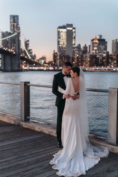 a bride and groom standing on a pier in front of the city skyline at night