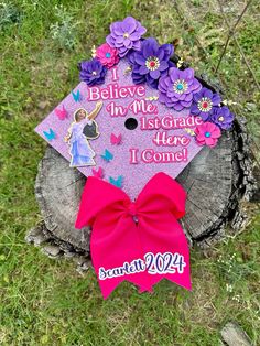 a purple graduation cap decorated with flowers and a pink ribbon on top of a tree stump