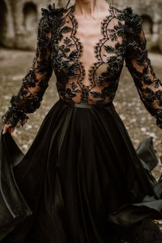 a woman wearing a black dress with sheer sleeves and flowers on the back, standing in front of an old building