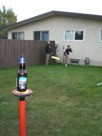 two men playing frisbee in the back yard with one man holding a beer